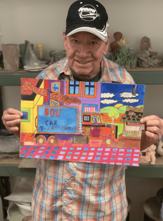 (An artist holding his artwork and smiling. He is an elder white man wearing a black baseball cap and a plaid shirt, with blue, orange and light orange colors. He is holding a drawing he made of a vibrantly colored box car.)