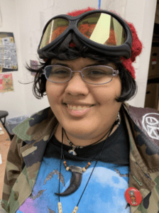 (The artist is a young Latinx woman wearing metal framed glasses and leather strung necklaces of various lengths. She is wearing ski goggles on the top of her head, a red knit beanie and a camo jacket)
