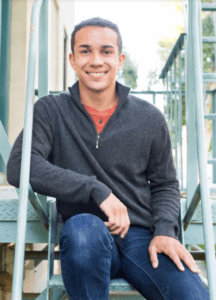 (The actor is young African American man, smiling at the camera, wearing a charcoal gray quarter-zip sweater, reddish-orange shirt underneath and blue jeans)